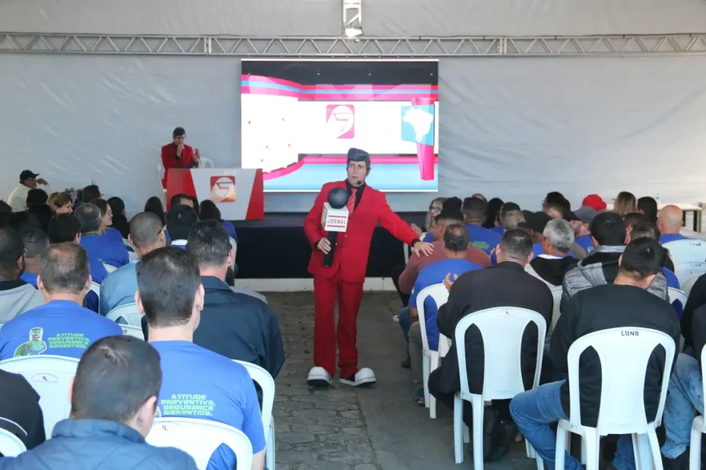 Personagem de palestra motivacional em terno vermelho e sapatos grandes, segurando um microfone "Jornal", interage com a plateia em evento de Segurança do Trabalho. Foco em Engenheiro de Segurança e Técnico de Segurança.