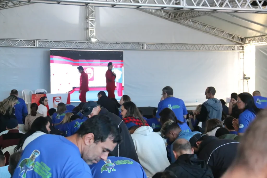 Dois palestrantes em ternos vermelhos no palco, em frente a um telão de LED, conduzem um evento de Segurança do Trabalho. A grande plateia, composta por funcionários com camisetas azuis de slogans de prevenção, participa do treinamento para Engenheiro e Técnico de Segurança.