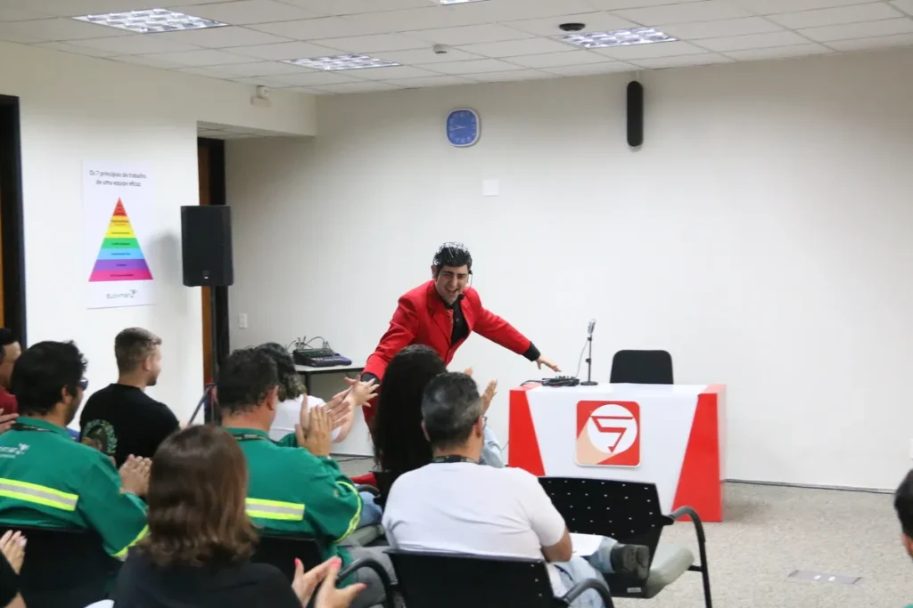 Palco com palestrante de terno vermelho sendo aplaudido pela plateia, incluindo participantes com uniformes, ilustrando o sucesso de uma palestra sobre exaustão mental e a conscientização na empresa.