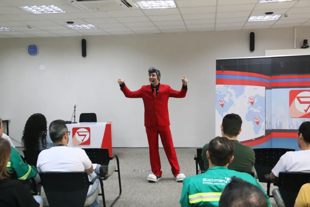 Palestrante com terno vermelho e sapatos brancos, fazendo um gesto enérgico em um palco, chamando a atenção de um auditório para a importância da palestra sobre exaustão mental e saúde no trabalho.