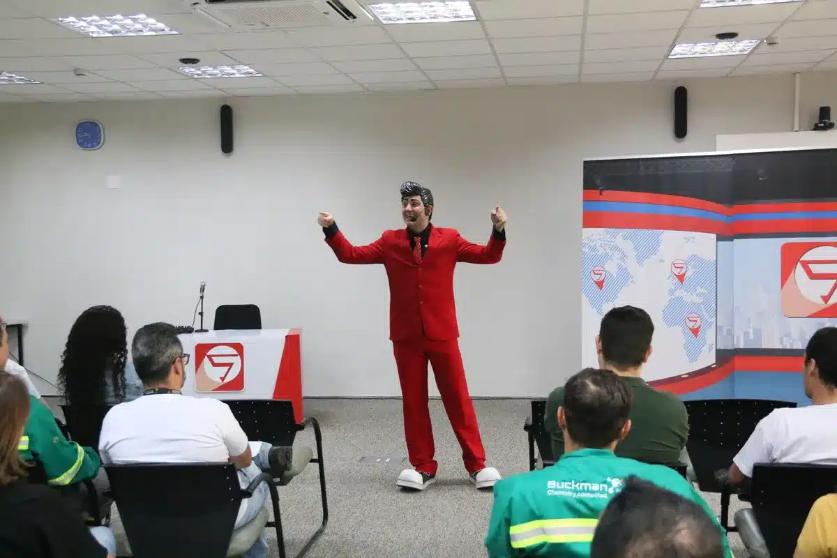 Palestrante com terno vermelho e sapatos brancos, fazendo um gesto enérgico em um palco, chamando a atenção de um auditório para a importância da palestra sobre exaustão mental e saúde no trabalho.