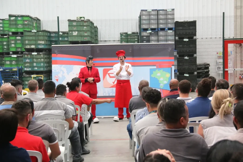 Dois palestrantes fantasiados em um evento corporativo. A plateia está sentada de frente para o palco, onde um apresentador vestido de chef e outro com terno vermelho e penteado se apresentam. O fundo mostra um galpão com caixas.