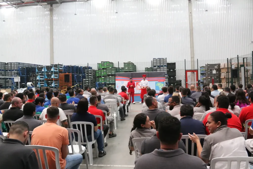 Vista ampla de uma plateia em um evento corporativo. Dois palestrantes fantasiados, um de terno vermelho e outro de chef, se apresentam para o público em um galpão.