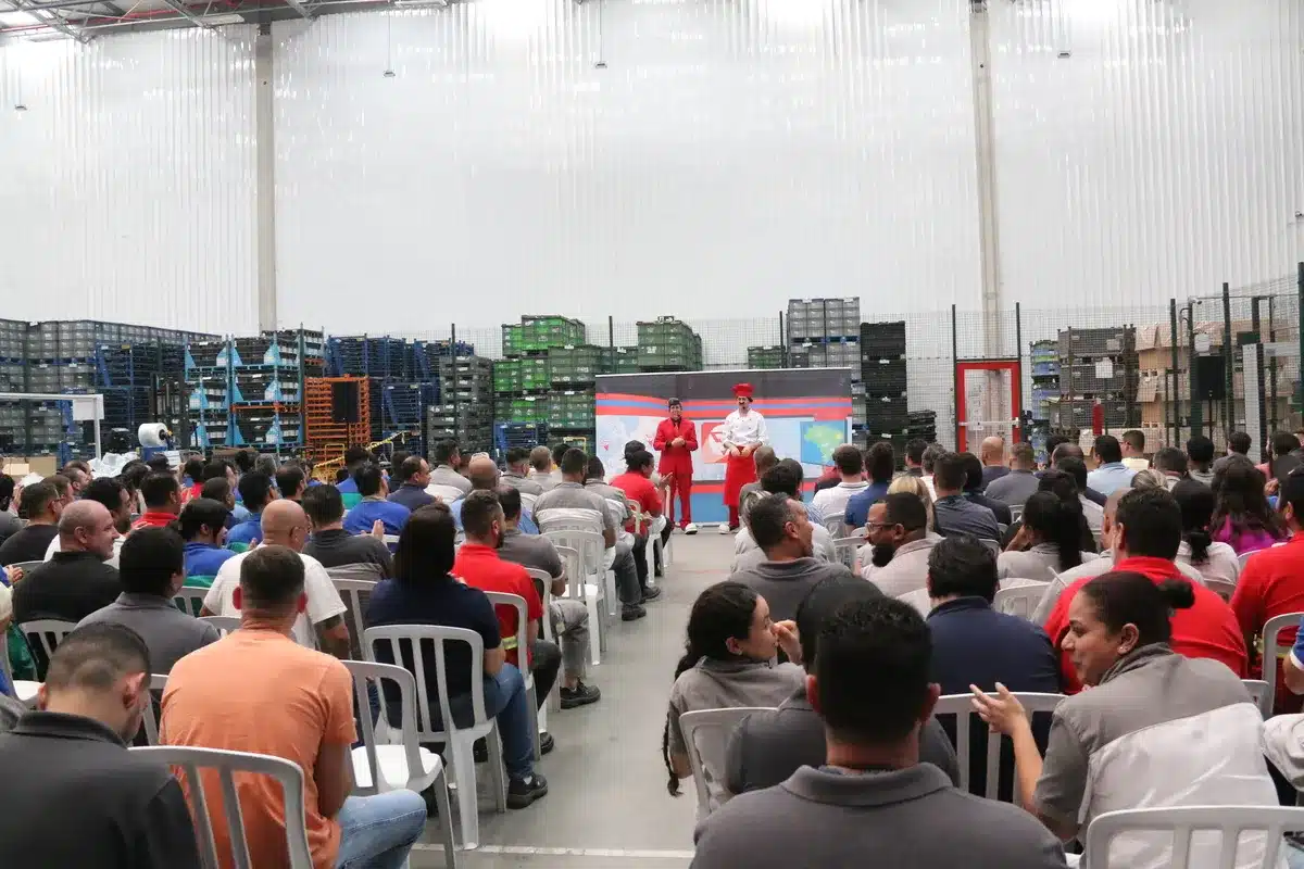 Vista ampla de uma plateia em um evento corporativo. Dois palestrantes fantasiados, um de terno vermelho e outro de chef, se apresentam para o público em um galpão.
