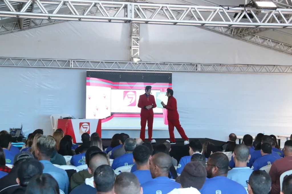 Dois palestrantes de fato vermelho a falar num palco em frente a uma grande plateia de colaboradores numa convenção de empresa, representando a entrega de frases motivacionais no trabalho.