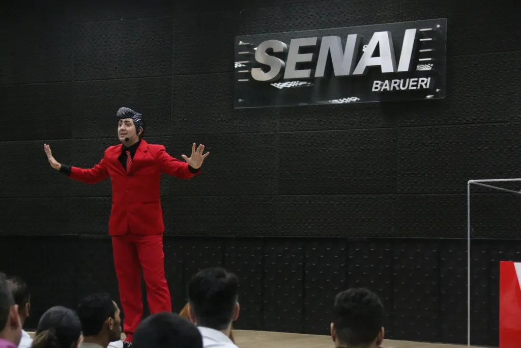 Palestrante de terno vermelho gesticula para a plateia durante evento motivacional em auditório do SENAI Barueri.