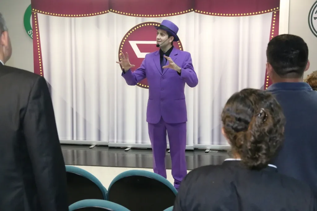 Um palestrante a gesticular num palco, com um fato roxo e chapéu, a falar para uma audiência em cadeiras azuis. A imagem simboliza a entrega de frases motivacionais curtas no ambiente de trabalho.