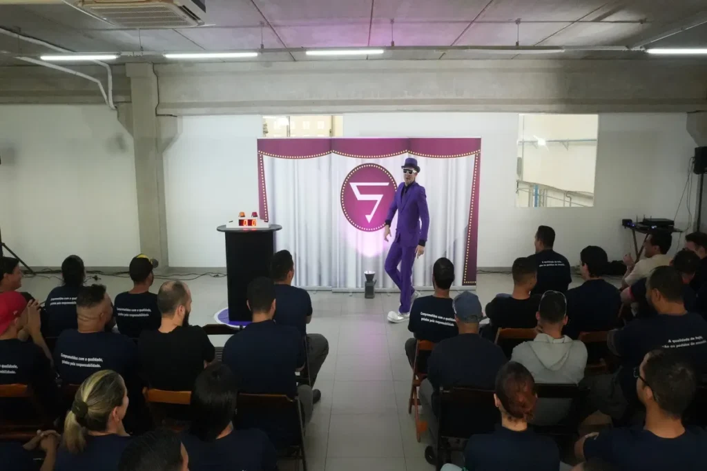 Palestrante de terno roxo e chapéu no palco, gesticulando para a plateia de colaboradores em evento motivacional ou SIPAT.