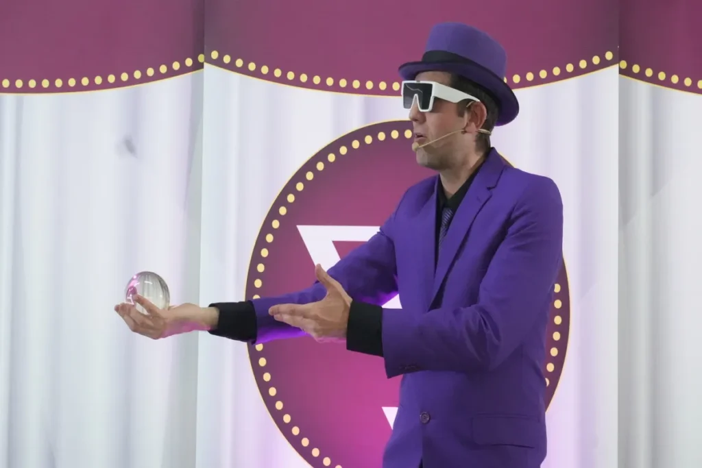 Close-up de um performista ou mestre de cerimônias no palco, vestido com um terno roxo, chapéu e óculos de sol futuristas, segurando uma bola de cristal transparente na palma da mão estendida. Ele está envolvido em uma apresentação lúdica, associada a eventos de palestra ginástica laboral ou SIPAT. O fundo tem cortinas brancas e um círculo magenta com um logo estilizado.