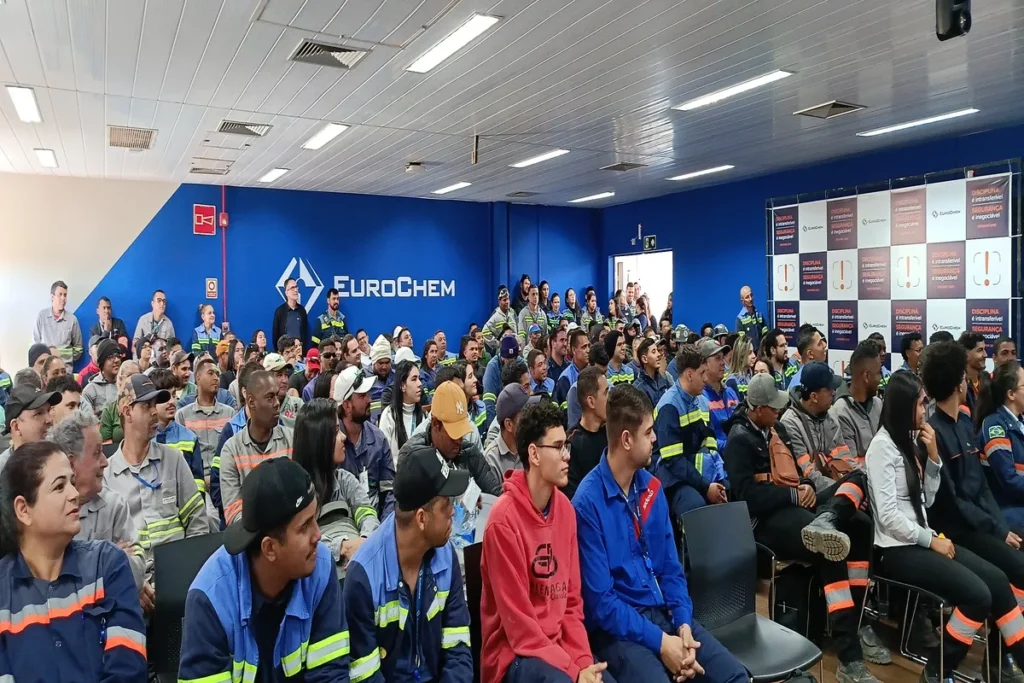 Multidão de funcionários de uma empresa em uniformes de trabalho assistindo a uma palestra em um auditório. A imagem representa uma reunião corporativa ou treinamento.