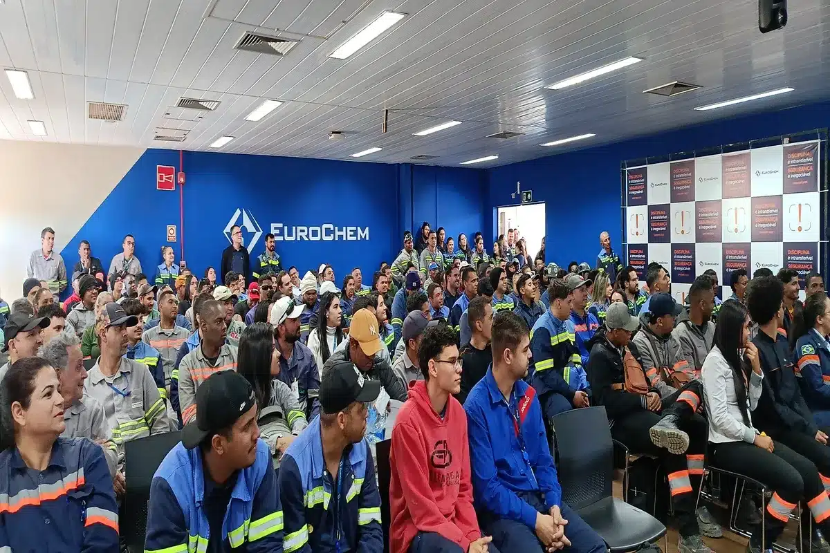 Multidão de funcionários de uma empresa em uniformes de trabalho assistindo a uma palestra em um auditório. A imagem representa uma reunião corporativa ou treinamento.