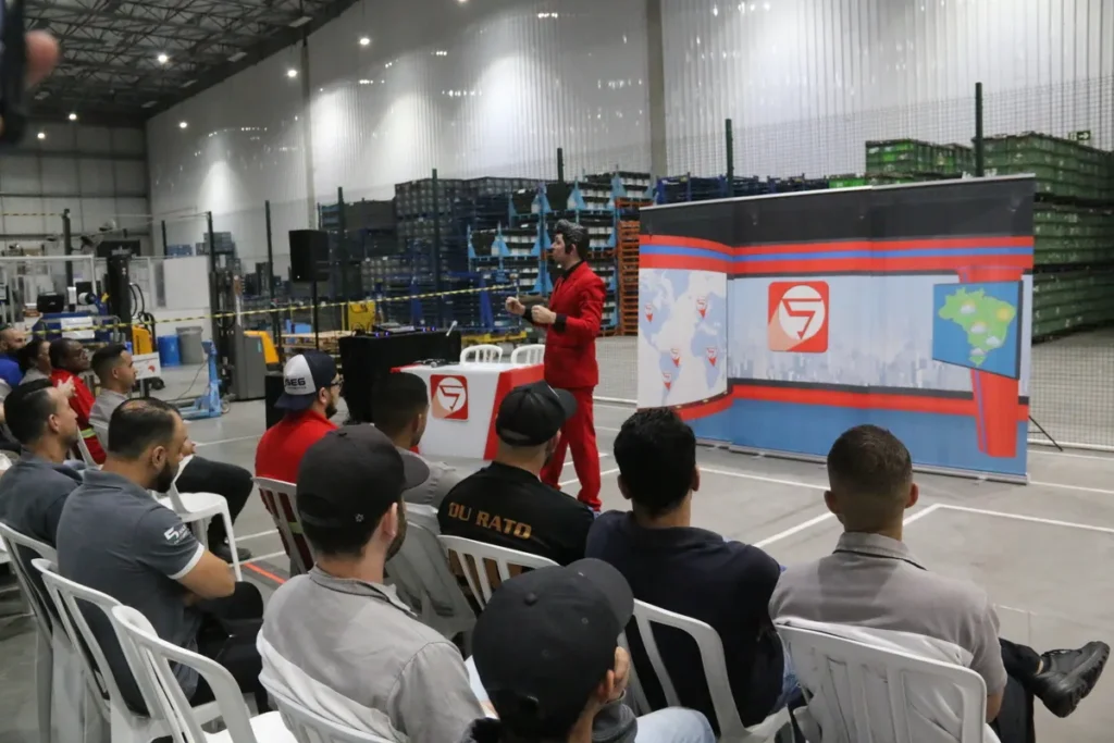 Um palestrante, vestido de vermelho e preto (como um repórter de segurança), faz uma apresentação para um grupo de funcionários em um galpão industrial com prateleiras ao fundo. O evento, que utiliza um painel temático de noticiário, provavelmente aborda temas de Segurança do Trabalho (SST) e higiene, cruciais para a prevenção da hantavirose e outras doenças transmitidas por roedores em ambientes de armazenamento e depósitos.