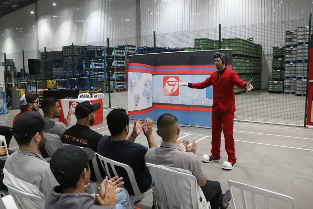 Palestrante em terno vermelho e preto se dirige a um público de operários em um galpão industrial, com prateleiras de estoque ao fundo, em um evento de Segurança do Trabalho. A palestra, com cenário de noticiário, foca em temas de higiene e prevenção de doenças transmitidas por roedores, como a Hantavirose, comum em áreas de depósito.