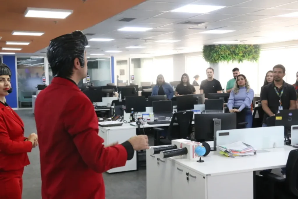 Duas pessoas vestidas de vermelho, uma de frente para a plateia e outra de costas, em uma apresentação. O homem, de costas, está de terno, enquanto a mulher, de lado, está com um terninho.