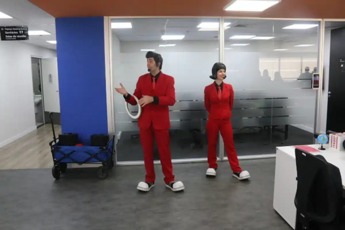 Duas pessoas vestidas de vermelho, uma de frente para a plateia e outra de costas, em uma apresentação. O homem, de costas, está de terno, enquanto a mulher, de lado, está com um terninho.