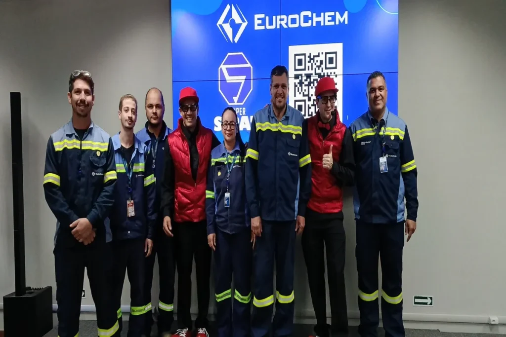 Grupo de seis homens e uma mulher em pé em um palco, vestindo uniformes de trabalho cinza e azul com faixas refletivas (EuroChem) e coletes vermelhos de instrutores, posando para uma foto durante um treinamento prático de segurança e emergências, como o manejo de hemorragia nasal.