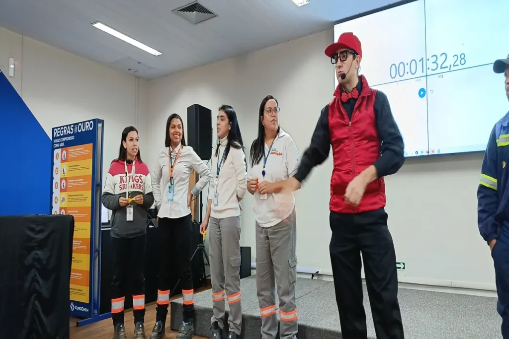 Treinamento de primeiros socorros em ambiente corporativo, mostrando quatro mulheres e um homem (instrutor de colete vermelho) em um palco. Um cronômetro digital no fundo marca a atividade em tempo real. O evento simula uma emergência como a hemorragia nasal para a equipe de brigada de incêndio ou CIPA.
