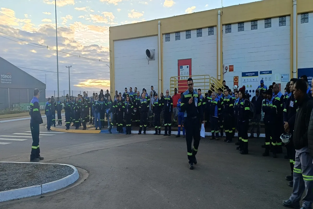 Grupo de funcionários de uma empresa, a maioria vestindo uniformes de trabalho com faixas refletivas azuis e amarelas, reunidos ao ar livre em frente a um galpão industrial, no contexto de um briefing ou treinamento de segurança e primeiros socorros.