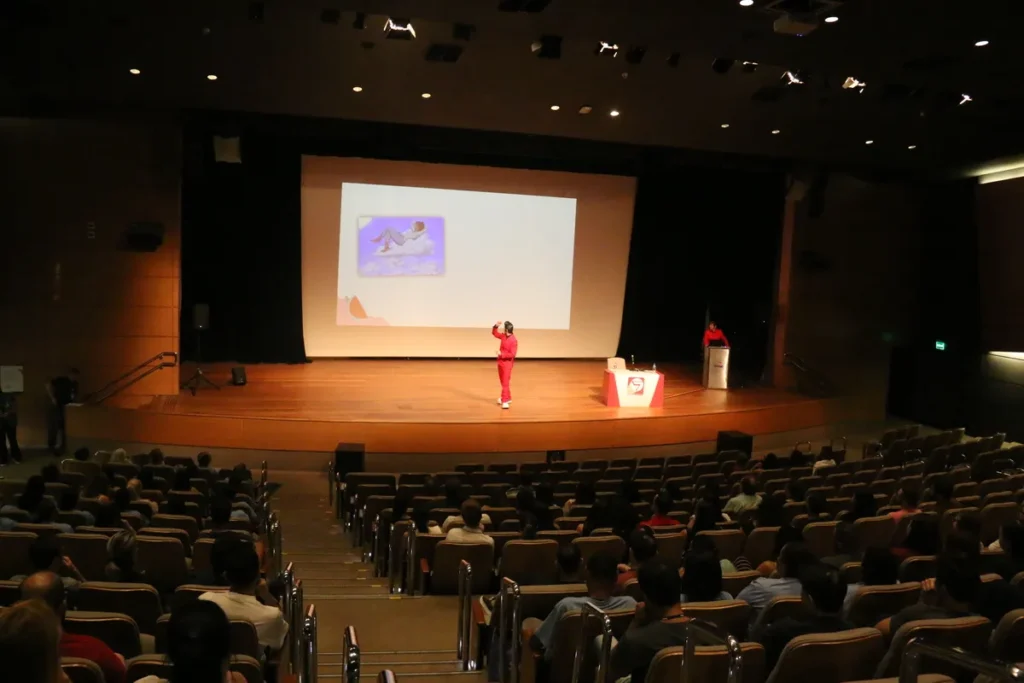 Uma pessoa, vestindo vermelho, em um palco de auditório. Ela gesticula em direção a um telão com uma ilustração. A cena pode representar uma palestra sobre saúde e bem-estar, abordando temas como a prevenção e tratamento da infecção fúngica.