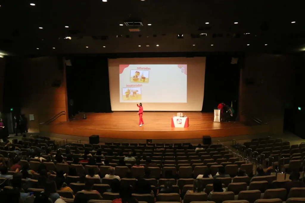 Uma pessoa com terno vermelho apresenta um slide sobre gorduras saturadas e insaturadas em um auditório cheio. A imagem pode ser associada a uma palestra sobre como a dieta e a nutrição podem influenciar na prevenção de doenças, como a infecção fúngica.