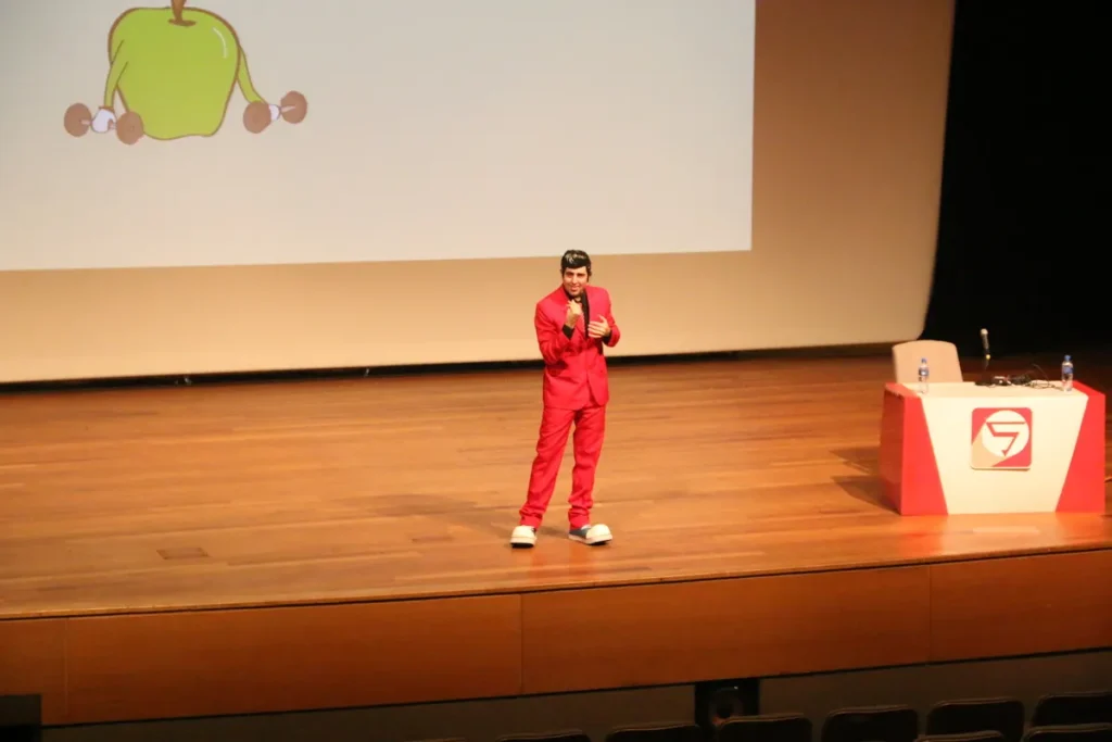 Um artista vestido com um terno vermelho e sapatos de palhaço está em um palco, segurando um microfone e com uma ilustração de uma maçã em um telão. A imagem pode ser usada para ilustrar uma palestra sobre a importância da alimentação e nutrição para prevenir doenças como a infecção fúngica.