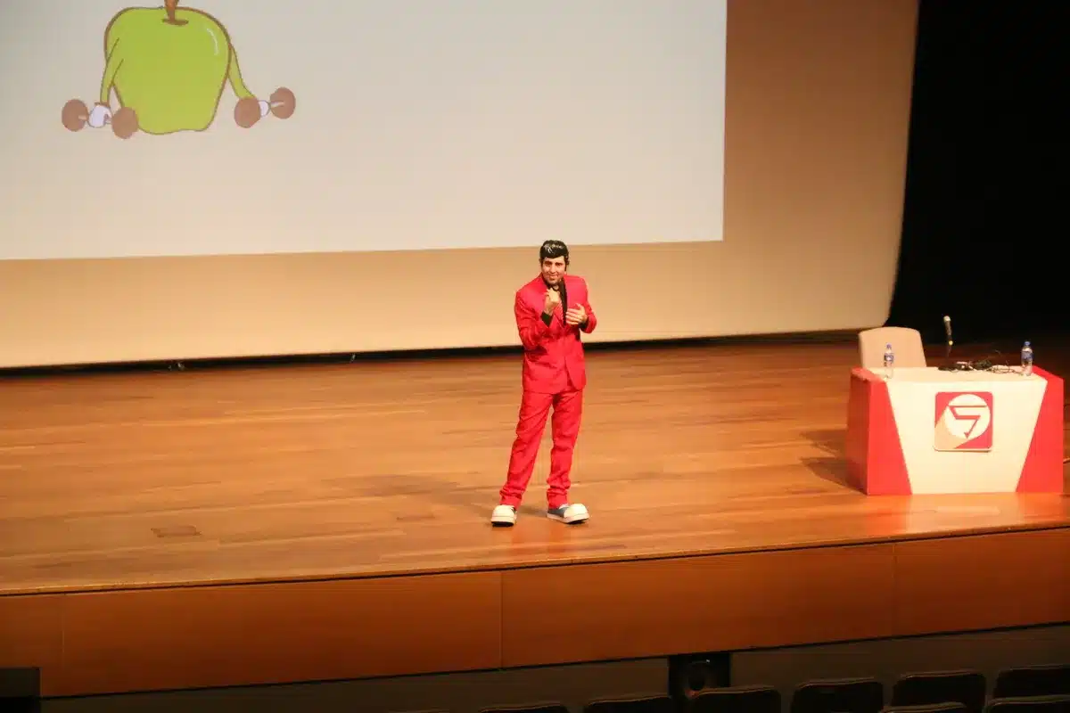 Um artista vestido com um terno vermelho e sapatos de palhaço está em um palco, segurando um microfone e com uma ilustração de uma maçã em um telão. A imagem pode ser usada para ilustrar uma palestra sobre a importância da alimentação e nutrição para prevenir doenças como a infecção fúngica.