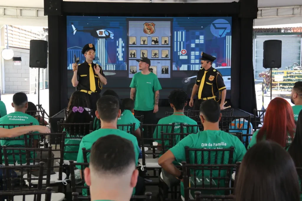inflamação joelho, dois homens em uniformes de segurança e um homem de camiseta verde em um palco, com um grupo de pessoas em cadeiras assistindo a uma palestra.