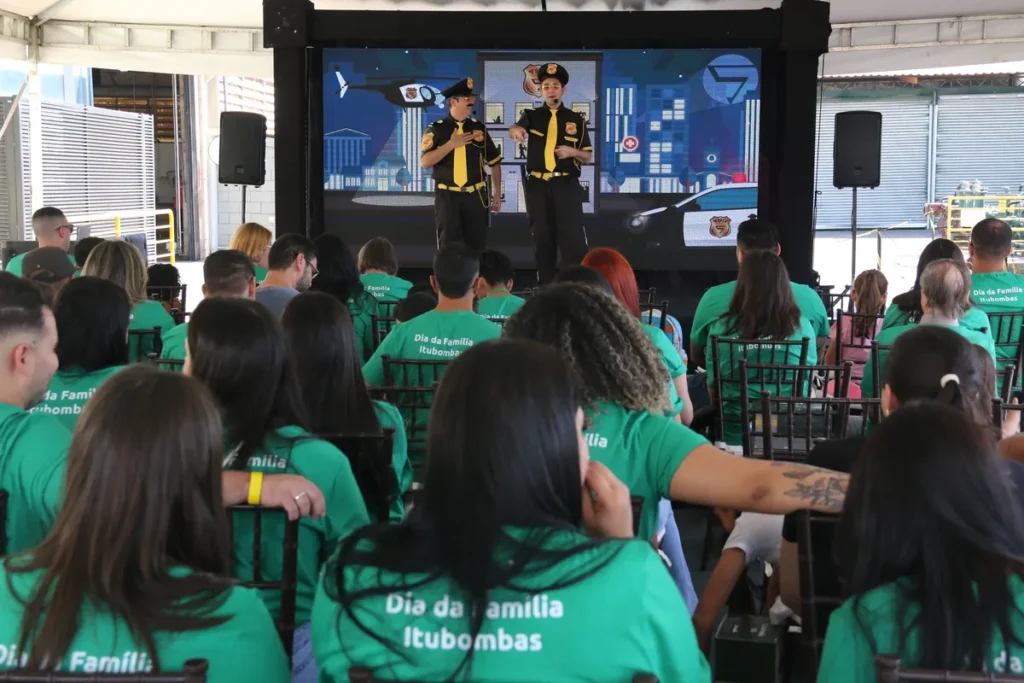 inflamação joelho, um grupo de pessoas em cadeiras, usando camisetas verdes com o logo "Dia da Família", assistindo a um evento com dois homens em um palco, com um telão exibindo um desenho animado de policial.