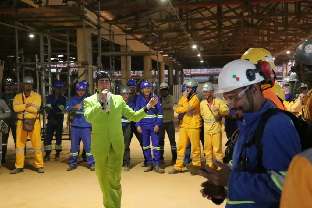 influenza, um homem em um terno verde-limão, segurando um microfone, de frente para um grupo de trabalhadores da construção civil.