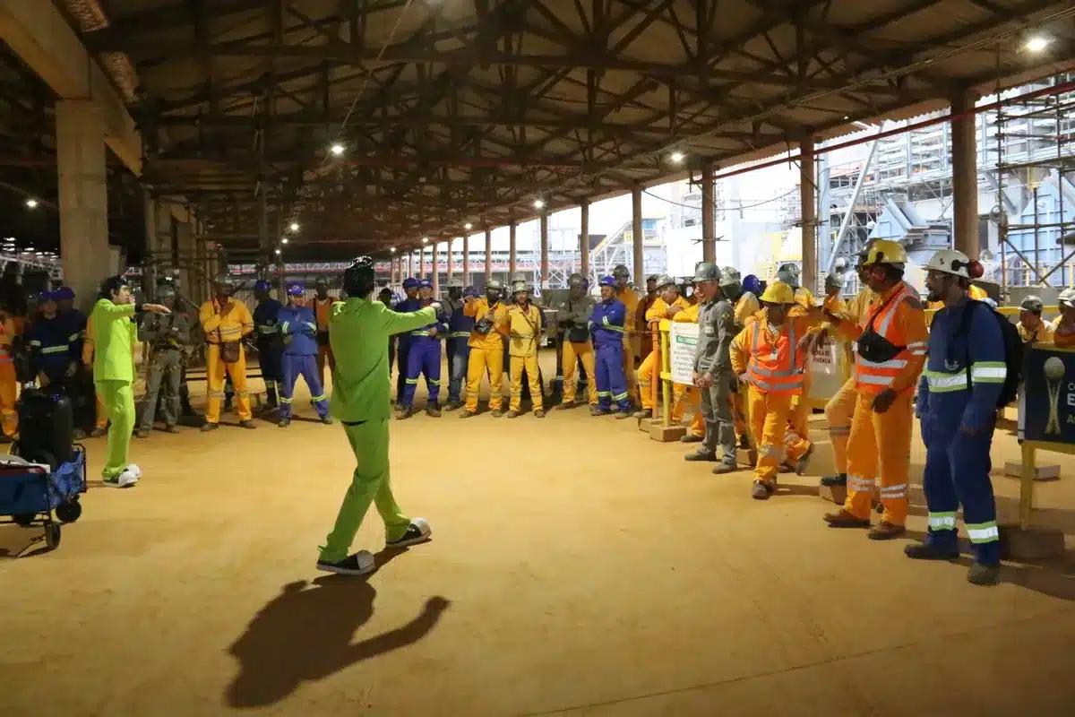 influenza, dois homens em ternos verde-limão, um deles com sapatos de Elvis Presley, de frente para um grupo de trabalhadores em um galpão.