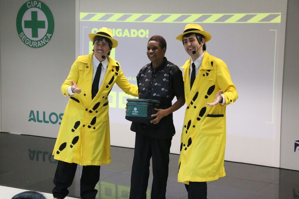 Dois apresentadores caracterizados com figurino amarelo realizam dinâmica sobre segurança do trabalho ao lado de colaboradora durante evento corporativo interno