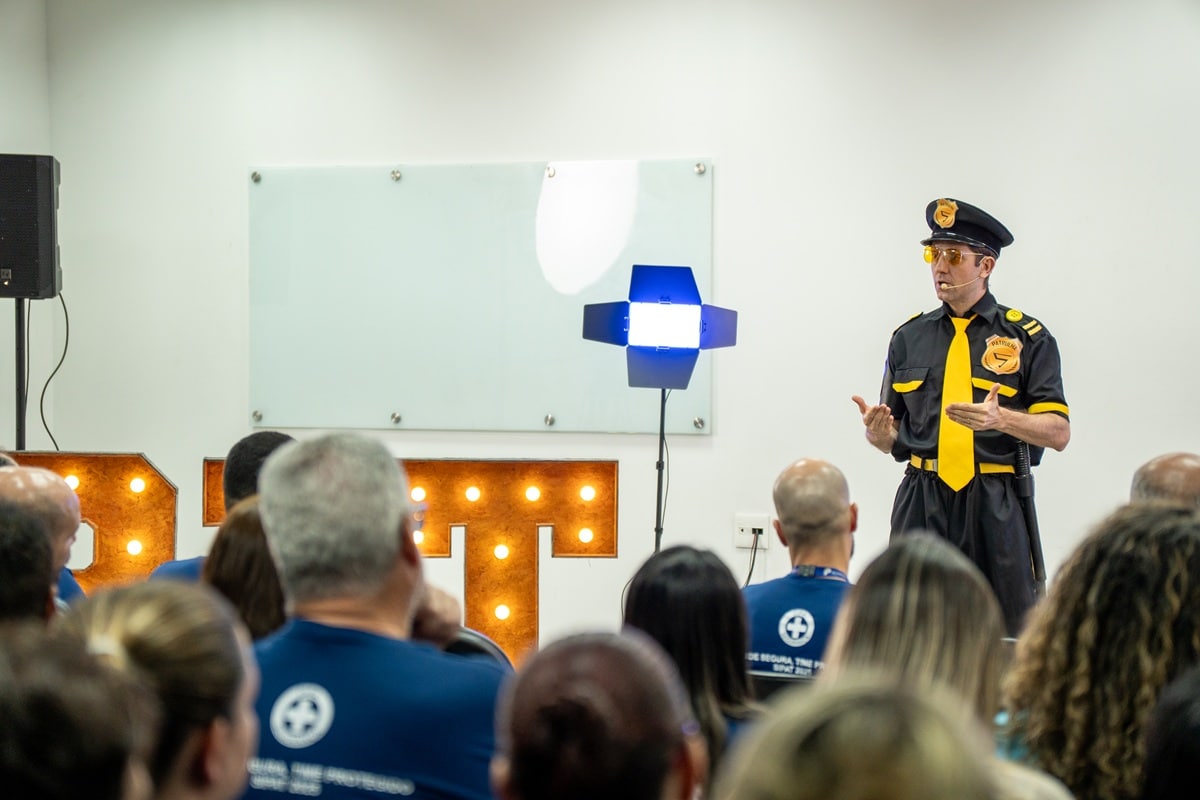 Palestrante vestido como agente de segurança, com uniforme preto e amarelo, falando em pé para um público sentado durante uma palestra corporativa em ambiente fechado