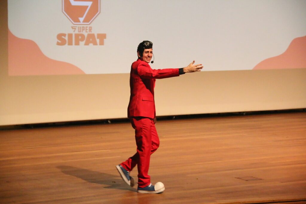 Palestrante de terno vermelho se apresenta no palco durante evento da SIPAT em auditório.