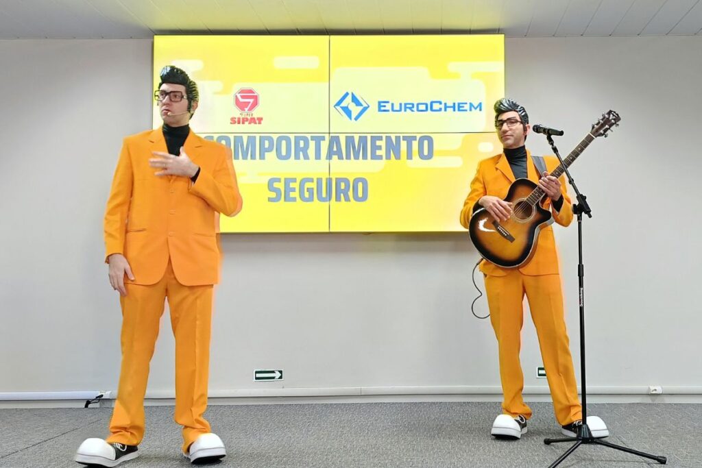 Apresentadores em trajes laranja realizam palestra show sobre comportamento seguro durante evento SIPAT corporativo.