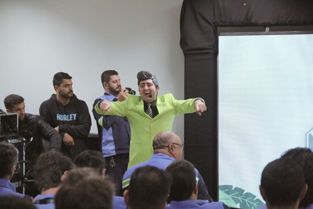 Palestrante com terno verde interagindo com público durante apresentação de SIPAT em ambiente corporativo.