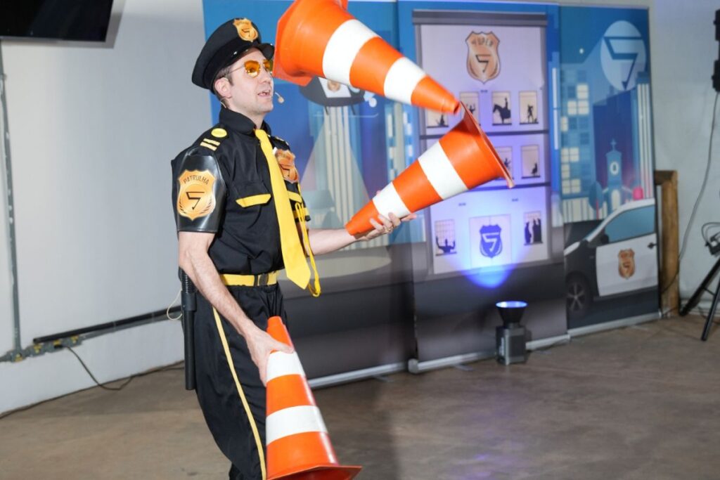 Palestrante caracterizado segurando cones laranja durante dinâmica em palestra gravada para SIPAT online.