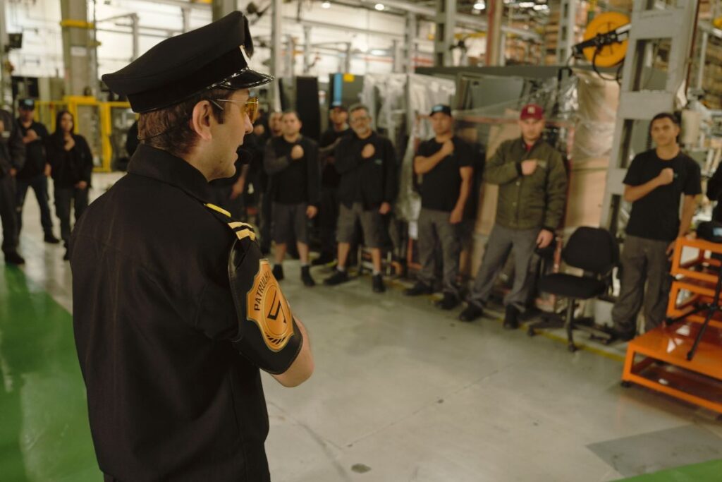 Palestrante caracterizado como agente de segurança fala com colaboradores reunidos em área industrial durante treinamento de SIPAT.