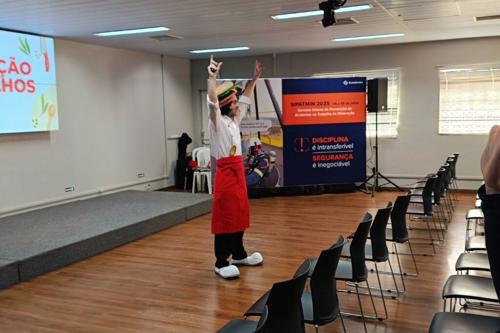Palestrante caracterizado conduzindo dinâmica em sala corporativa com cadeiras e banner de SIPAT ao fundo.