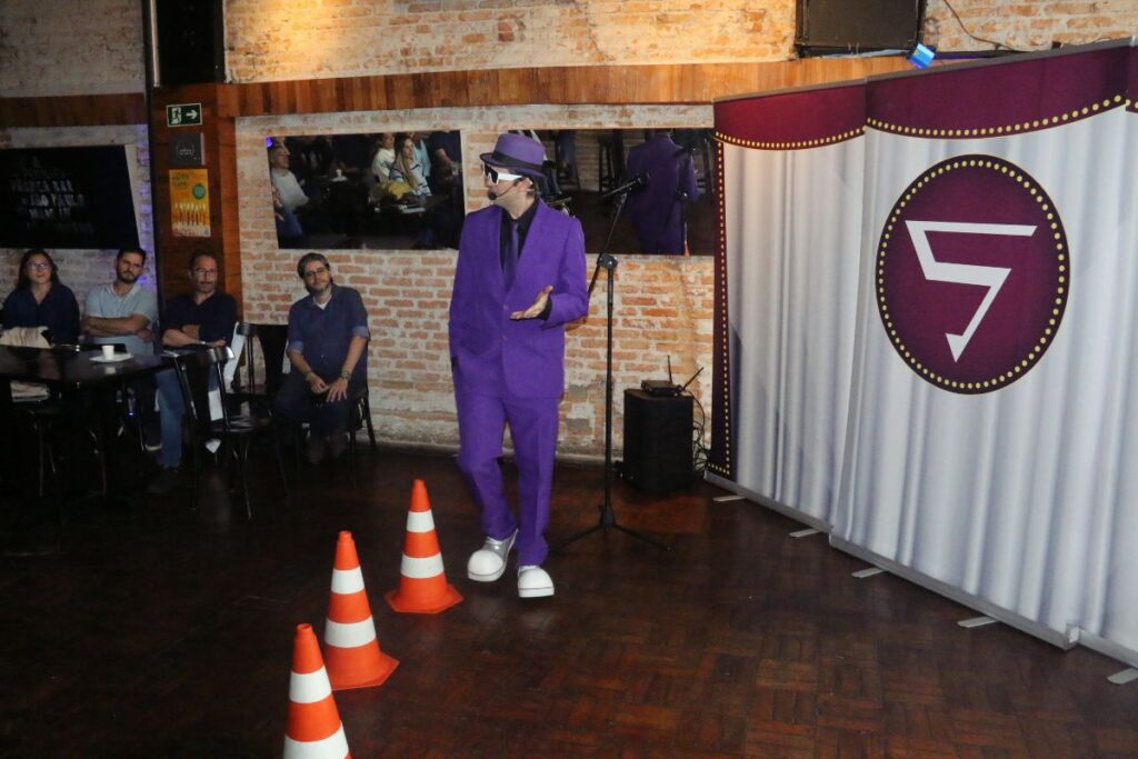 Personagem vestido de roxo conduz palestra interativa de SIPAT em ambiente fechado com plateia e cones de sinalização no palco.