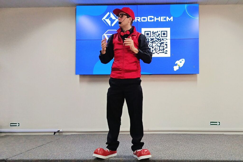 Palestrante caracterizado com roupa vermelha conduzindo apresentação interativa para SIPAT online em frente a uma tela digital.