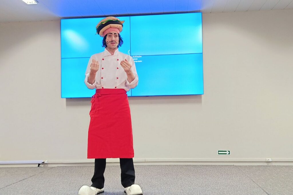 Palestrante fantasiado de chef conduzindo apresentação de SIPAT online em frente a uma tela digital.
