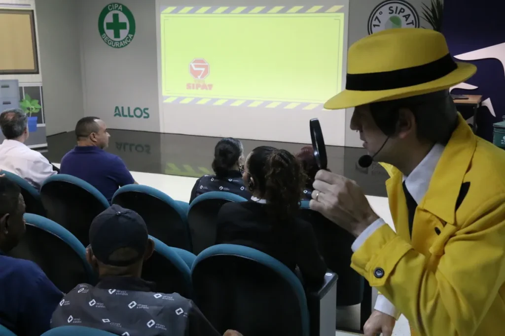 Close-up de palestrante vestido como detetive, usando chapéu e casaco amarelo, segurando uma lupa e um microfone, durante uma palestra sobre intoxicação alimentar e segurança (SIPAT).
