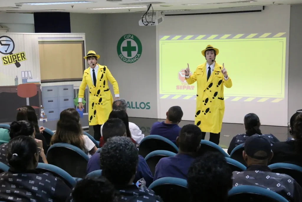 Dois palestrantes vestidos como detetives de fantasia amarela, gesticulando no palco para uma plateia atenta, em uma palestra sobre intoxicação alimentar e segurança (CIPA/SIPAT).