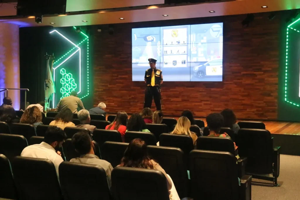 Um mestre de cerimônias fantasiado de policial ou agente de segurança apresenta para uma plateia atenta sentada em um auditório. O telão ao fundo exibe uma grade de imagens e gráficos relacionados à segurança, incluindo o logo CIPA e ilustrações de prevenção de acidentes. O ambiente é moderno, com paredes de madeira e iluminação de LED verde. Uma bandeira com um emblema fica visível à esquerda do palco.