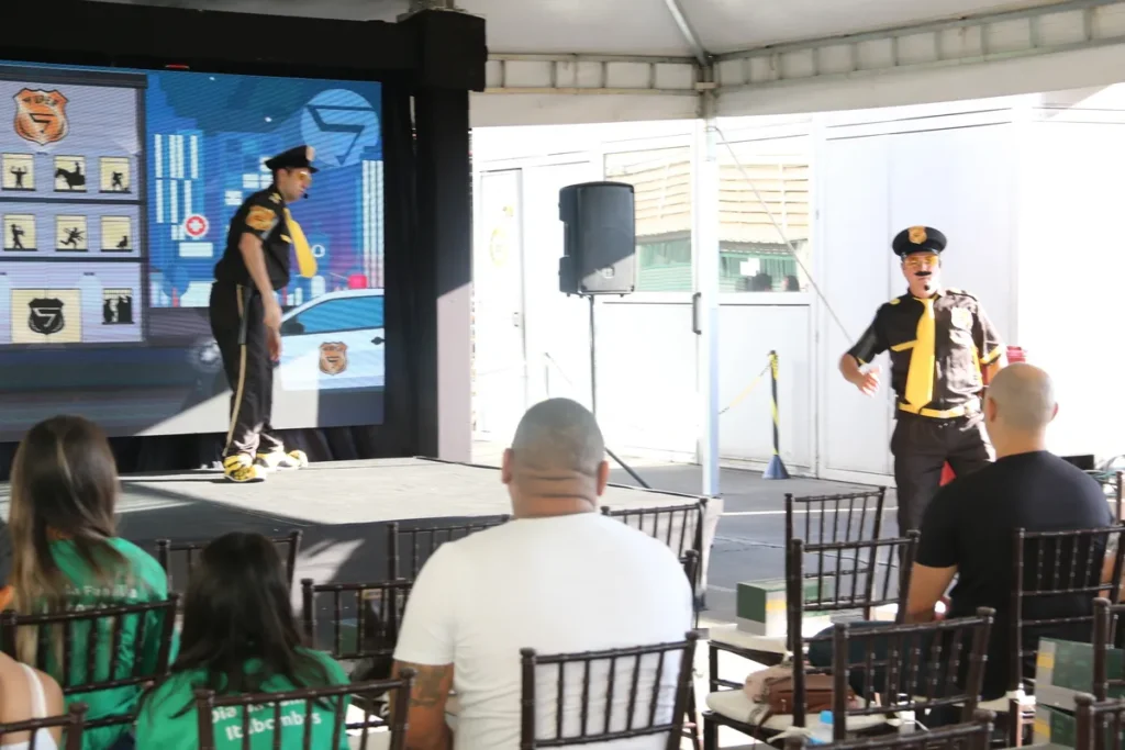 Dois atores ou palestrantes vestidos com fantasias de agentes de segurança (uniforme preto e gravata amarela) em um palco, interagindo com o público de um evento ao ar livre (provavelmente um "Dia da Família"). O telão ao fundo exibe gráficos de segurança do trabalho. A cena é de uma apresentação lúdica focada em prevenção de doenças ocupacionais, como a lombalgia.