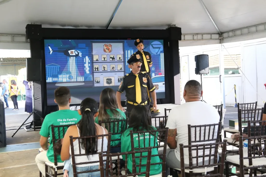 Dois palestrantes ou atores performando no palco, vestidos como personagens de segurança (uniforme preto e amarelo), em um evento ao ar livre chamado "Dia da Família Itubombas". O telão exibe ilustrações de segurança do trabalho (incluindo o logo SIPAT) e o público usa camisetas verdes, associadas à empresa. A cena é de uma ação de conscientização lúdica sobre segurança e saúde, frequentemente incluindo a prevenção de lombalgia.