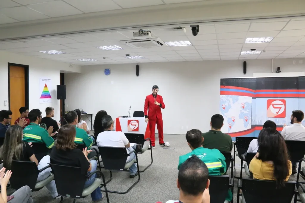 Palestrante com terno vermelho em uma palestra de segurança sobre Mapa de Risco. Ele interage com a plateia de funcionários uniformizados e casuais em uma sala de treinamento, com um telão exibindo gráficos de segurança.