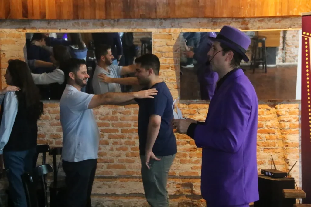 Um artista de terno roxo e chapéu interage com duas pessoas, que estão praticando exercícios de equilíbrio e coordenação em uma sala com paredes de tijolos. A imagem pode ser usada para ilustrar workshops de saúde ou fisioterapia para o tratamento de condições como a neuropatia periférica.
