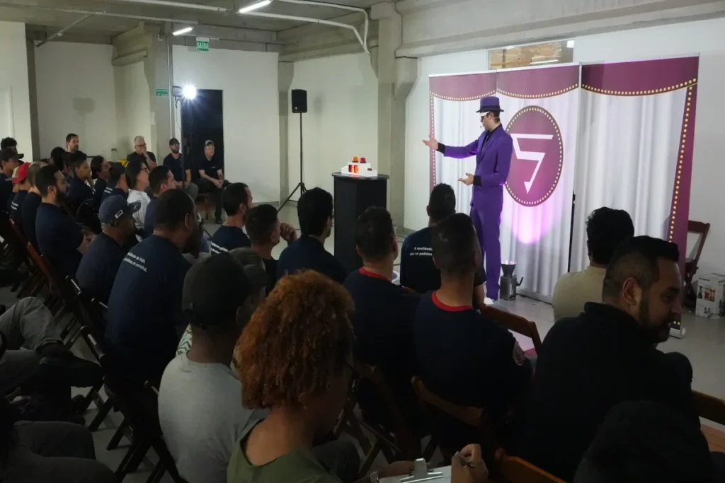 Um palestrante com um terno roxo e chapéu se dirige a um auditório com trabalhadores, durante uma apresentação sobre a norma regulamentadora NR 13.