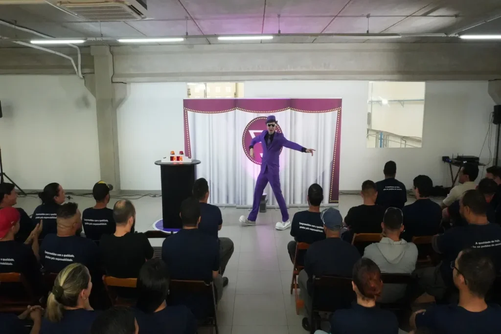 Um palestrante em terno roxo, óculos de sol e chapéu, se apresenta de forma teatral em um palco para um público sentado, durante uma palestra sobre a NR 13.